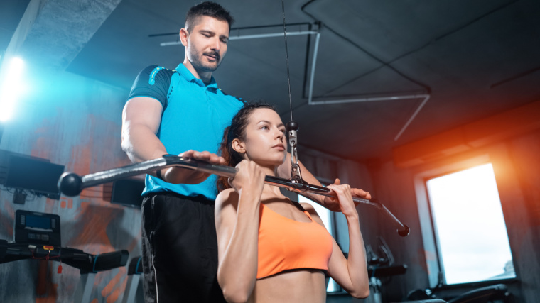 An athlete performs a reverse-grip lat pulldown with support from their coach.