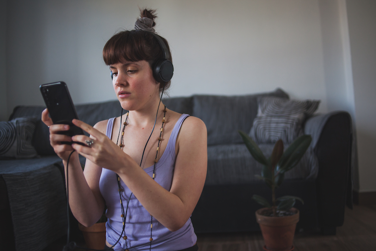 A woman with headphones sitting on the floor in front of a couch looking at a smartphone during an at-home workout.